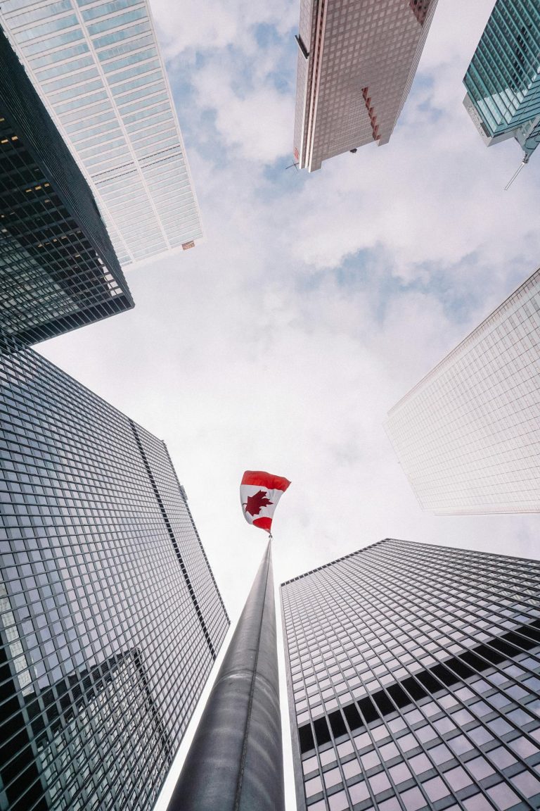 From below of perspective tunnel between glass skyscrapers and pole with fluttering flag of Canada